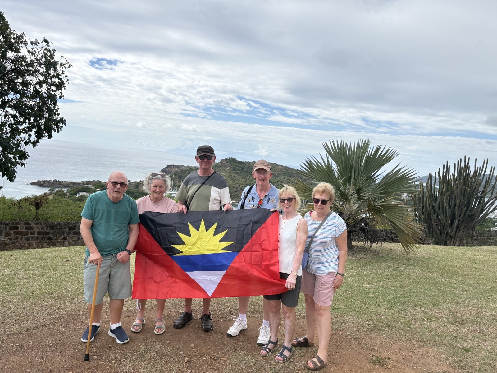 Six guests holding the Antigua and Barbuda flag at a scenic coastal lookout.